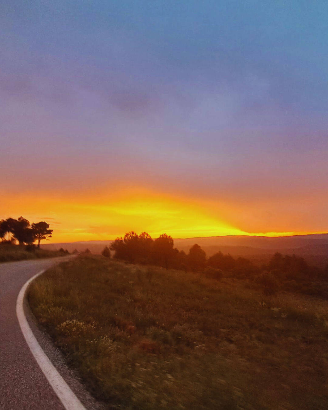 Landstraße führt in Richtung eines intensiven Sonnenuntergangs mit orangefarbenem Himmel und dunklen Baumumrissen.